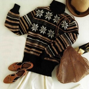 Peut inclure: Un pull rayé noir et marron avec un motif flocon de neige, un pantalon blanc, un sac cabas en maille marron et des chaussures en cuir marron.