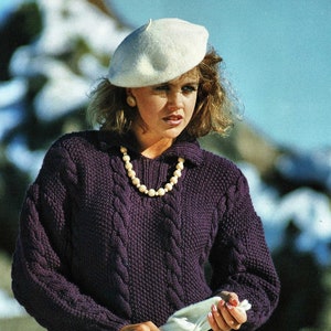 May include: A woman wearing a dark purple cable-knit sweater with a white beret and pearl necklace. The sweater has a collared neckline and long sleeves. She is holding a white cloth. The background is blurred.