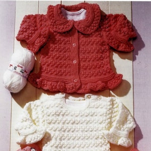 May include: Two knitted baby sweaters with ruffled edges. The top sweater is pink with a white collar and buttons. The bottom sweater is white with long sleeves. Both sweaters are made with a textured stitch.