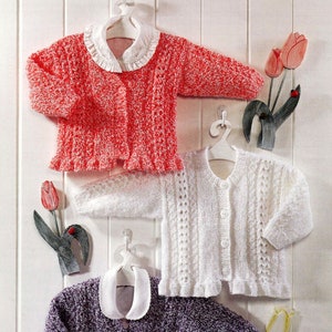 May include: Three knitted baby cardigans in pink, white and purple with white collars and button fastenings. The cardigans are hanging on hangers against a white wood panel background.