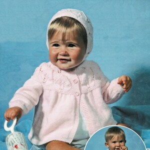 May include: A pink knitted baby cardigan with a button closure and a white crocheted bonnet. The cardigan has a delicate lace pattern and is worn by a baby sitting on a blue surface.