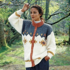 Op de afbeelding: Een wit, crème en blauwe gebreide cardigan met een geometrisch patroon en een knoopsluiting. De cardigan heeft een hoge kraag en lange mouwen.