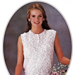 May include: A white sleeveless knitted top with a button closure at the neck. The top has a textured, bobbly knit pattern. The model is wearing the top with a floral skirt and holding a bouquet of flowers.