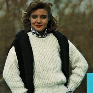 Könnte beinhalten: Ein weißer Strickpullover mit gerippter Struktur. Der Pullover hat einen runden Ausschnitt und lange Ärmel. Das Model trägt ein kariertes Hemd unter dem Pullover.