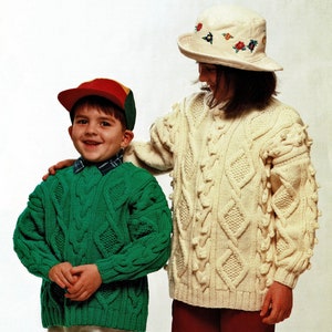 Puede incluir: Dos niños con suéteres de punto. La niña lleva un suéter blanco con un patrón de punto de cable y un sombrero blanco con flores bordadas. El niño lleva un suéter verde con un patrón de punto de cable y una gorra de béisbol roja, amarilla y verde.