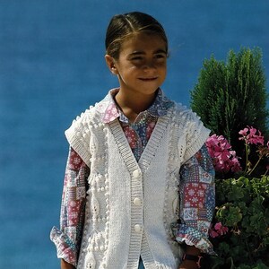 May include: A young person wearing a white knitted waistcoat with a button fastening, a patterned shirt, and blue trousers. The waistcoat has a textured design with small white bobbles.