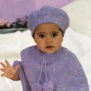 May include: A baby wearing a purple knitted poncho with pom-poms and a matching beret. The poncho and beret have a speckled pattern with multi-colored flecks.