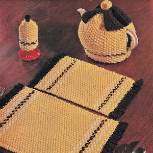 Puede incluir: Dos manteles individuales de ganchillo amarillo y negro con bordes con flecos y un cubre-tetera de ganchillo con la parte superior negra.