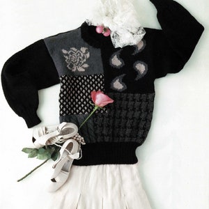 Puede incluir: Un suéter negro y gris de patchwork con un cuello de encaje blanco y adornos de flores rosas. El suéter se combina con una falda blanca y zapatos blancos.