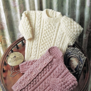 Puede incluir: Dos suéteres de punto para bebé, uno blanco y otro rosa, con un patrón de trenza. Los suéteres están colocados sobre una mesa de madera.