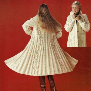 May include: A long, white knitted coat with a cable knit pattern. The coat has a high collar and a button closure. The model is wearing the coat with brown boots.