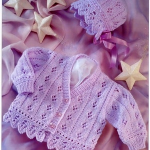 May include: A lavender knitted baby cardigan with a delicate lace pattern and button closure. The cardigan is laid on a purple fabric background with decorative stars.