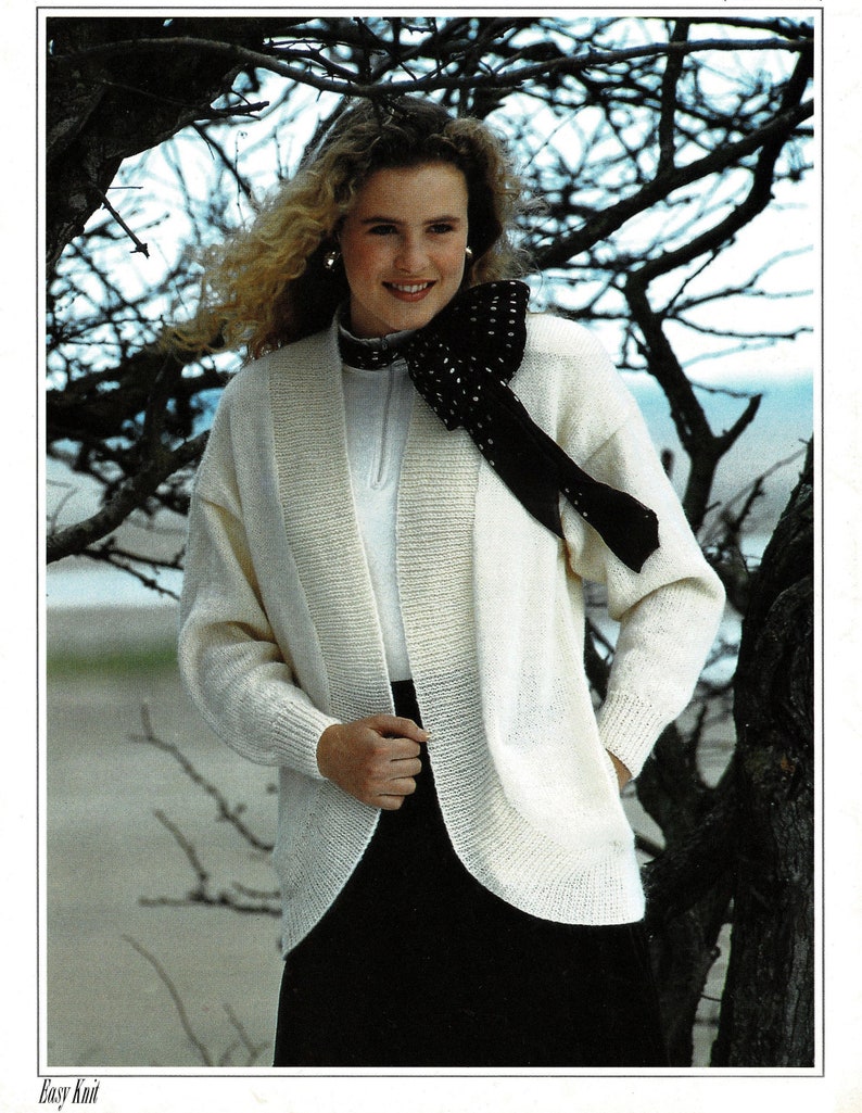 May include: A white knitted cardigan with a shawl collar and a black and white polka dot scarf tied around the neck. The cardigan has long sleeves and a relaxed fit. The model is standing in front of a tree with bare branches.