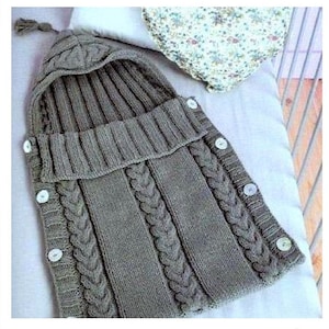 May include: A gray knitted baby sleeping bag with a hood and buttons. The sleeping bag has a cable knit pattern and a white tassel.