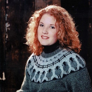 May include: A woman with red curly hair is wearing a grey sweater with a white and grey sheep pattern. The sweater has a high neck.