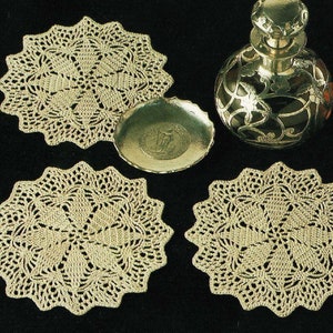 Puede incluir: Tres doilies de ganchillo en color beige, con un diseño decorativo, se muestran sobre una superficie negra. También se muestra en la imagen una botella de plata con un diseño decorativo.
