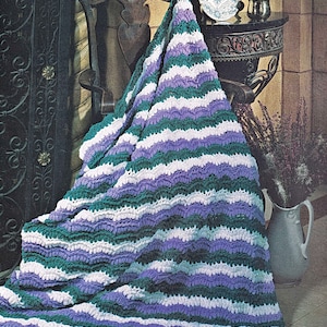 May include: A crocheted blanket with a wavy pattern in shades of purple, green, and white. The blanket is draped over a wooden chair and a table.