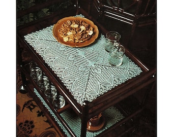 May include: A close-up of a knitted lace trolley cloth on a dark wooden trolley. The cloth is light blue and has a delicate lace pattern. The trolley is decorated with a brown patterned rug.
