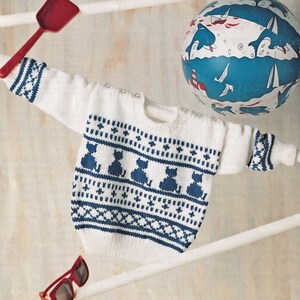 Peut inclure: Un pull en tricot blanc avec un motif de chats bleu et blanc. Le pull a un col rond et des manches longues.