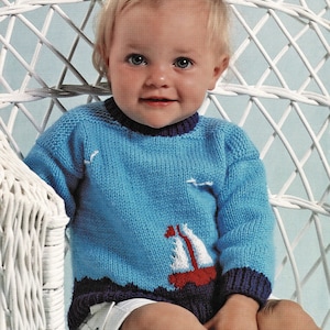 Può includere: Un bambino che indossa un maglione blu lavorato a maglia con un design di barca a vela rossa e bianca. Il maglione ha un bordo blu scuro intorno al collo e alle maniche. Il bambino è seduto su una sedia di vimini bianca. DOPPIO TRICOT 46-56 cm 18-22 pollici