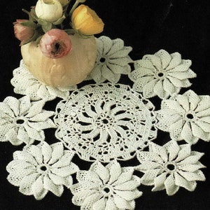 Könnte beinhalten: Eine Anordnung aus weißen, gehäkelten Deckchen in verschiedenen Blumenformen. Ein zentrales kreisförmiges Deckchen ist von blütenförmigen Teilen umgeben. Eine kleine Vase mit rosa und gelben Blumen sorgt für einen Farbtupfer und schafft ein Vintage-Feeling.