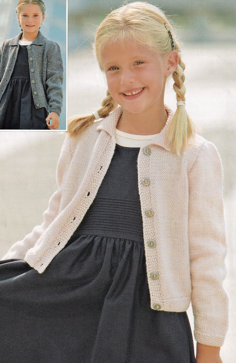 May include: A young girl wearing a light pink knitted cardigan with a collared neckline and button closure. She is wearing a black dress underneath the cardigan. The cardigan has a textured knit pattern and is buttoned up the front.