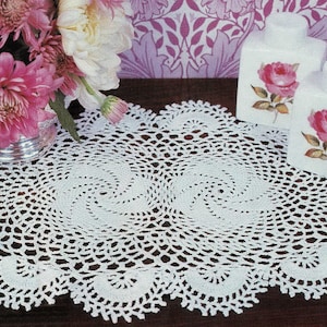 Può includere: Una tovaglietta ovale bianca all'uncinetto con bordo smerlato e motivo a spirale. Due barattoli di ceramica bianca con illustrazioni di rose a destra. Un mazzo di fiori rosa e bianchi in un vaso d'argento.