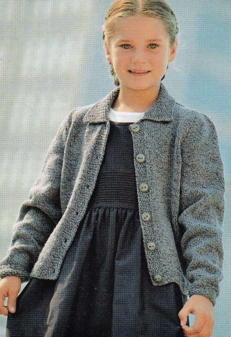 May include: A young person wearing a gray knitted cardigan with a dark gray dress underneath. The cardigan has a button-up front and a collared neckline.