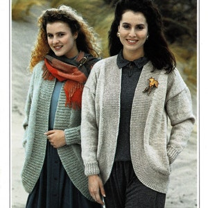 May include: Two women wearing knitted cardigans, one in a light blue cardigan with a red scarf and the other in a beige cardigan. Both cardigans have a relaxed fit and a slightly open front. The women are standing in a sandy outdoor setting.