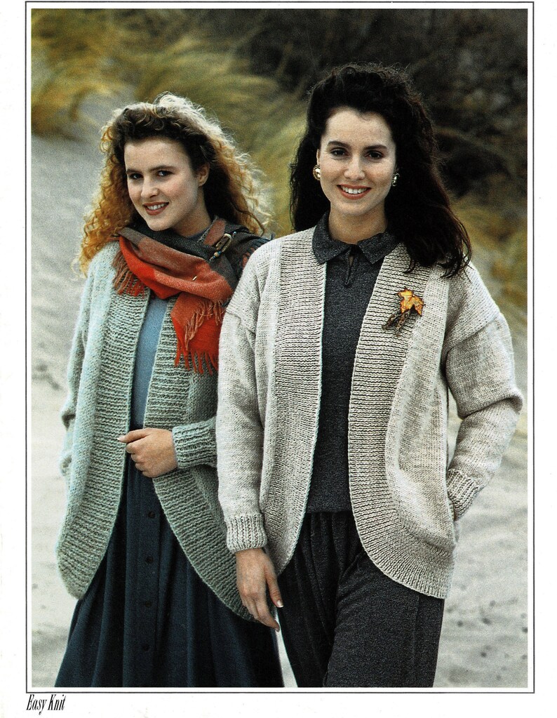 May include: Two women wearing knitted cardigans, one in a light blue cardigan with a red scarf and the other in a beige cardigan. Both cardigans have a relaxed fit and a slightly open front. The women are standing in a sandy outdoor setting.