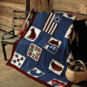 Puede incluir: Una colcha de ganchillo azul y roja con varios diseños cuadrados, incluyendo una bandera americana, un gato, una casa y una sandía. La colcha está sobre una silla de madera con un violín. Una cesta de mimbre llena de piñas también es visible.