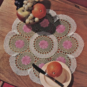 Könnte beinhalten: Eine weiße Häkeldeckchen mit grünen und rosa Akzenten zeigt ein florales Design. Die Deckchen liegt auf einem Holztisch mit einer Schüssel Obst und einem Teller mit einer Orange.