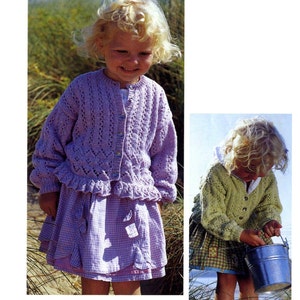 May include: Two young girls wearing knitted cardigans. The girl on the left is wearing a purple cardigan with a ruffled hem and a white and purple gingham skirt. The girl on the right is wearing a green cardigan and a plaid skirt.