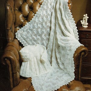 May include: A white, knitted baby blanket and matching dress are draped over a brown leather armchair. The blanket features a scalloped edge and a repeating leaf pattern. The dress has a round neckline and button closure.