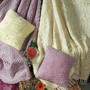 Può includere: Un'accogliente composizione di coperte e cuscini lavorati a maglia in morbidi colori pastello. Le coperte sono color crema e lavanda, con motivi e trame intricati. Due cuscini quadrati, uno color crema e uno lavanda, aumentano il comfort. Un cesto di vimini e fiori completano la scena.