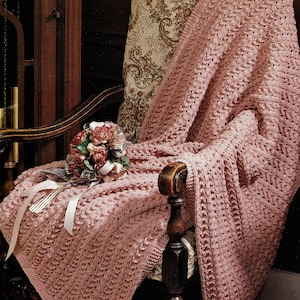 Może przedstawiać: Różowy pled szydełkowy drapowany na drewnianym fotelu z tapicerką w kwiatowy wzór. Pled ma fakturowany wzór ściegu i jest starannie złożony na fotelu.