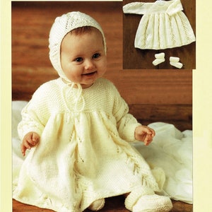 Puede incluir: Un conjunto de vestido, gorro y patucos de punto blanco para bebé. El vestido tiene un patrón de encaje y un lazo en el cuello. El gorro es de estilo boina con un borde de encaje. Los patucos son sencillos y tienen un puño abatible.