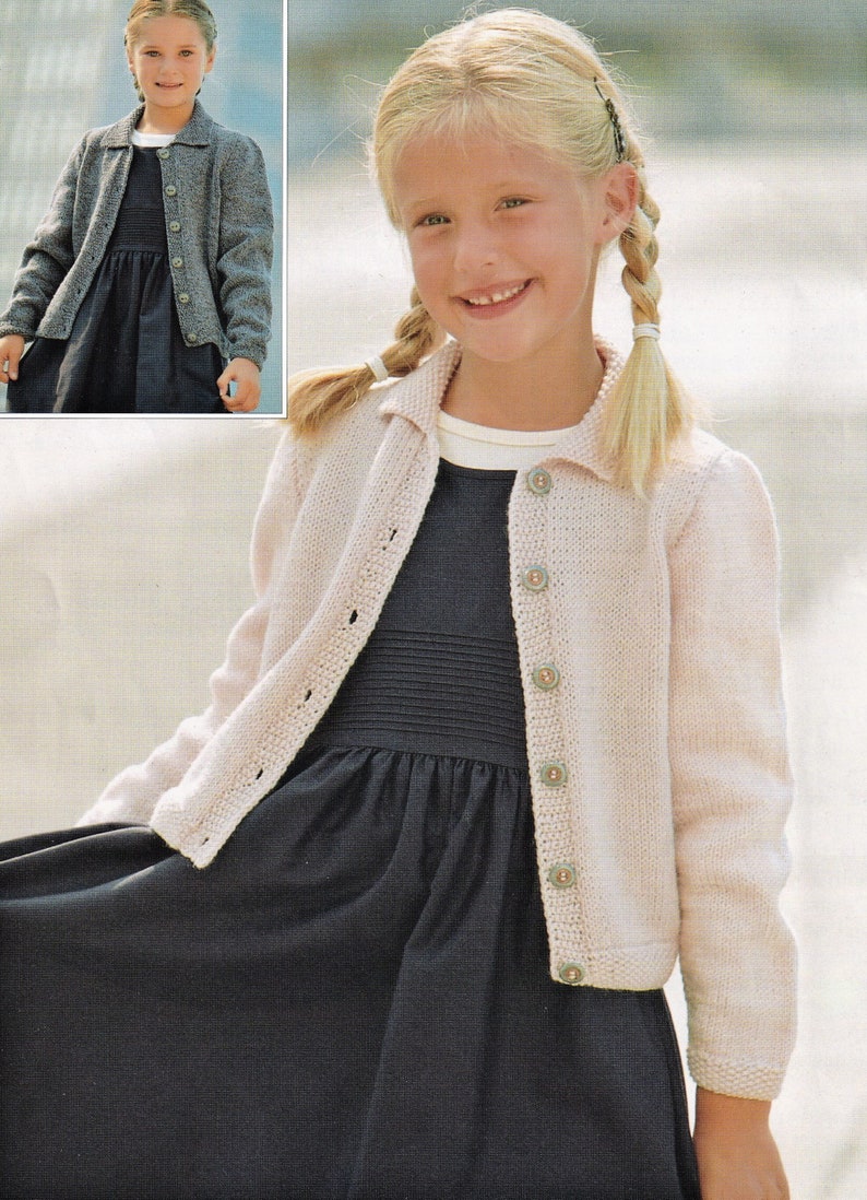 May include: A young girl wearing a light pink knitted cardigan with a collared neckline and button closure. She is wearing a dark blue dress underneath the cardigan. The cardigan has a delicate stitch pattern and is a classic style.