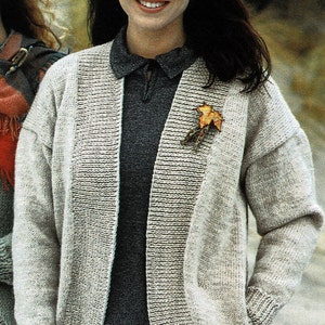 May include: A woman wearing a light grey cardigan with a dark grey shirt underneath. The cardigan has a long, open front and a leaf-shaped pin on the left side. The woman has long dark hair and is smiling.