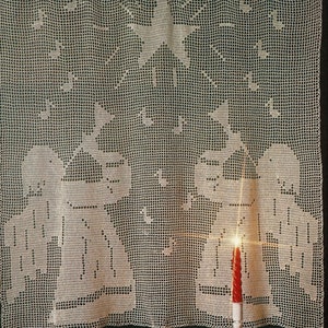 May include: White crocheted curtain with a star and a Christmas scene. The curtain is hanging on a wooden rod with rings. A red candle is lit in front of the curtain.