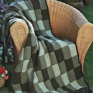 May include: A green and brown crocheted throw blanket with fringe, draped over a wicker chair. The blanket is made of squares in different shades of green and brown.