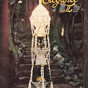 Peut inclure: Un porte-plante en macramé blanc à trois niveaux avec un abat-jour blanc en haut. Le porte-plante est décoré de perles jaunes et a des glands blancs en bas. Le texte "Macrame Elegance II" est écrit en lettres dorées en haut de l'image. Le texte "Basic to Advanced Designs Including Macrame Necklaces" est écrit en bas de l'image. Le texte "De Lune" est écrit en bas à gauche de l'image.