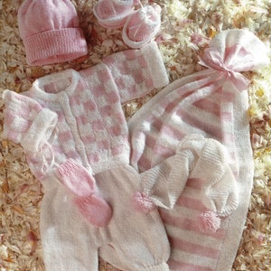 Puede incluir: Un conjunto de ropa y accesorios para bebé en rosa y blanco. El conjunto incluye un gorro de punto con pompón, patucos, una rebeca, un pelele, manoplas y una manta. La manta tiene un lazo rosa atado.