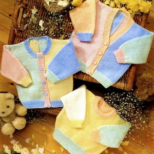 Puede incluir: Tres cárdigans de punto para bebé de colores diferentes, con combinaciones de colores rosa, azul, amarillo y blanco. Los cárdigans están colocados en una cesta de mimbre con flores y pétalos esparcidos alrededor.