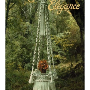 May include: A white macrame plant hanger with a glass plate and a pot of orange flowers hanging in a forest setting. The text "Macrame Elegance" is written in gold script above the hanger. The text "La Creation" is written in black below the hanger.