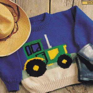 Peut inclure: Un pull bleu tricoté avec un motif de tracteur vert et jaune sur le devant. Le pull a un bas de couleur crème et un col rond. Un chapeau de paille et une botte noire sont visibles.