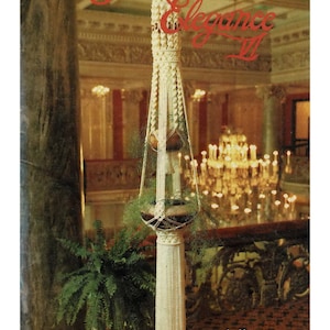 May include: A white macrame plant hanger with a tassel, hanging from the ceiling in a grand room with a chandelier. The text "Macrame Elegance II" is written in red at the top of the image. The text "Embellissement" is written in red at the bottom of the image.