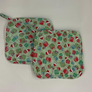 May include: Two square fabric pot holders featuring a light green background with a repeating pattern of red, green, and blue gift boxes, white snowflakes, candy canes, and red and white Santa hats. Each pot holder has a small fabric hanging loop.
