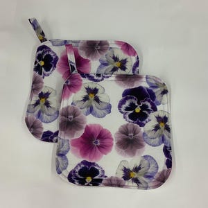 May include: Two square fabric pot holders featuring a repeating floral pattern of pansies in shades of purple, violet, magenta, and white. Each quilted kitchen accessory includes a small fabric loop at the top corner for convenient hanging storage.