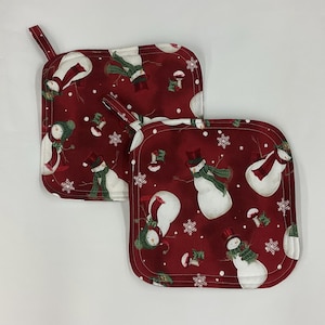 May include: Two square, red Christmas potholders with a snowman pattern. The snowmen have top hats, scarves, and carrot noses. White snowflakes and small green and red accents are scattered across the fabric. Each potholder has a small fabric loop.
